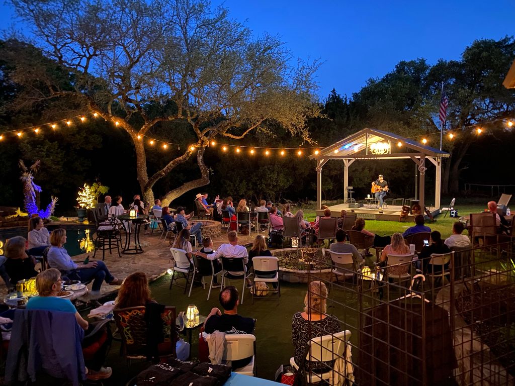 A full house at dusk: guests seated under the string lights, a songwriter alone on the gazebo stage